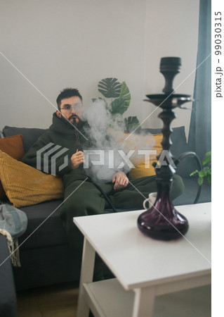 Bearded man is smoking hookah at home and blowing cloud of smoke - chill time and resting concept Bearded man is smoking hookah at home and blowing cloud of smoke - chill time and resting concept 99030315