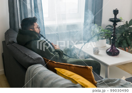 Bearded millennial or gen z man smoking hookah while relaxing on sofa at home - chill time and resting concept 99030490