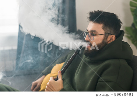 Bearded man is smoking hookah at home and blowing cloud of smoke - chill time and resting concept Bearded man is smoking hookah at home and blowing cloud of smoke - chill time and resting concept 99030491