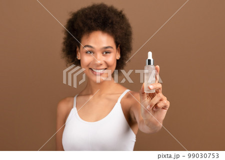 Smiling young african american curly female with perfect skin shows jar of serum recommends cosmetics 99030753