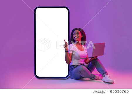 Online Offer. Happy black woman with laptop pointing at big blank smartphone Online Offer. Happy black woman with laptop pointing at big blank smartphone 99030769