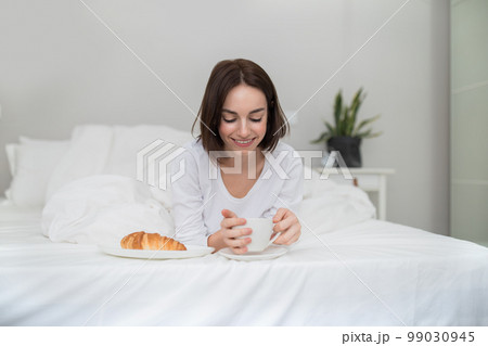 Happy relaxed young woman drinking coffee, eating pastry in bed Happy relaxed young woman drinking coffee, eating pastry in bed 99030945