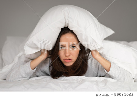 Tired sad caucasian millennial female lies on bed under white blanket, covers head with pillow, suffers from insomnia Tired sad caucasian millennial female lies on bed under white blanket, covers head with pillow, suffers from insomnia 99031032