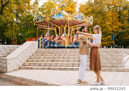 Wide shot of romantic young couple in love standing hugging looking at each other in amusement park in summer day.. Handsome bearded man and pretty blonde woman embracing enjoying weekend outdoors. 99032265