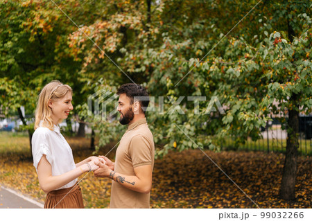Portrait of happy young couple in love standing at city park and looking at each other with falling in love eyes. Bearded handsome man and blonde woman walking spending time together on summer day. 99032266
