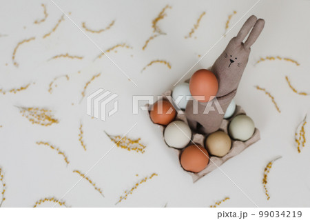 Funny handmade bunny rabbit toy with colorful chicken eggs and yellow mimosa flowers. Easter gift concept Funny handmade bunny rabbit toy with colorful chicken eggs and yellow mimosa flowers. Easter gift concept 99034219
