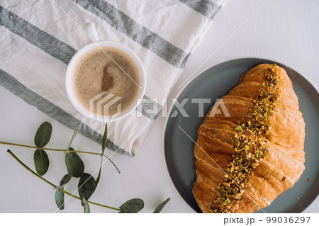 Flat lay of cup of cappuccino with kitchen towel, croissant and eucalyptus branch on the table Flat lay of cup of cappuccino with kitchen towel, croissant and eucalyptus branch on the table 99036297