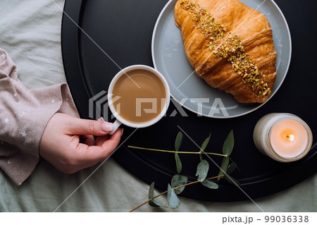 Flat lay of woman's hand holding cup of cappuccino, croissant and scented candle with eucalyptus branch on the tray Flat lay of woman's hand holding cup of cappuccino, croissant and scented candle with eucalyptus branch on the tray 99036338