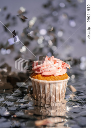 A cupcake with a pink cap stands among the flying silver confetti. 99044950