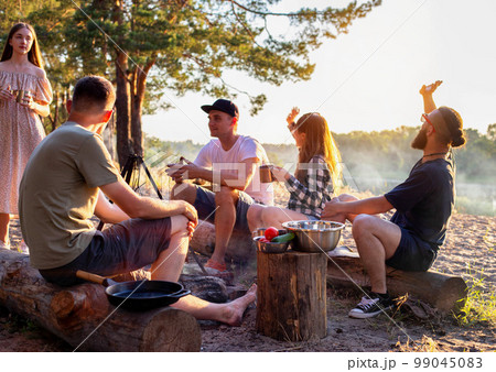A company of young people have a rest in nature in the summer against the backdrop of the forest and the river bank. Tourism and hiking in summer. Copy space for text 99045083
