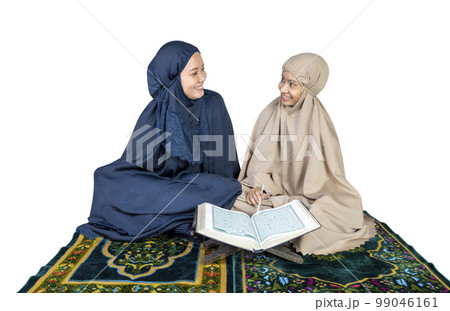Asian Muslim mother and daughter in a hijab on a prayer rug reading the Quran 99046161
