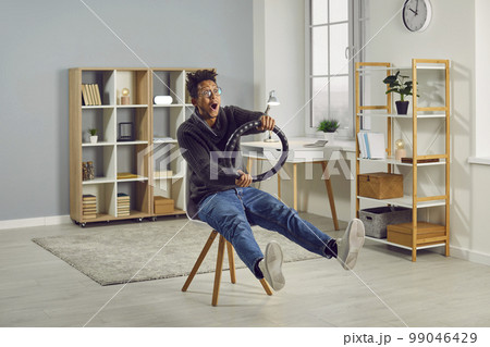Funny, crazy man pretending to drive an invisible car at a great speed on a chair at home 99046429