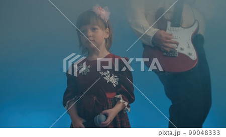 Little girl on stage in vintage dress, she sings into microphone and dances, her father plays an electric guitar. Color music is shining and smoke is billowing. Family time, creativity and hobbies Little girl on stage in vintage dress, she sings into microphone and dances, her father plays an electric guitar. Color music is shining and smoke is billowing. Family time, creativity and hobbies 99048333