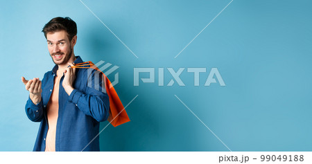 Modern guy holding shopping bag behind shoulder and beckon you, telling to come closer or inviting to store, standing on blue background Modern guy holding shopping bag behind shoulder and beckon you, telling to come closer or inviting to store, standing on blue background 99049188
