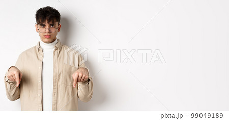 Portrait of handsome hipster guy in glasses showing an ad, pointing fingers down at logo, standing on white background 99049189