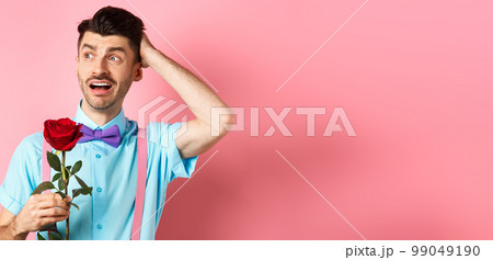 Nervous man waiting for his date on Valentines day, holding red rose and looking confused sideways, scratching head anxiously, standing on pink background 99049190