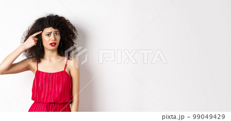 Shocked and confused caucasian woman in red dress scolding someone crazy, pointing finger at head and frowning, standing over white background 99049429