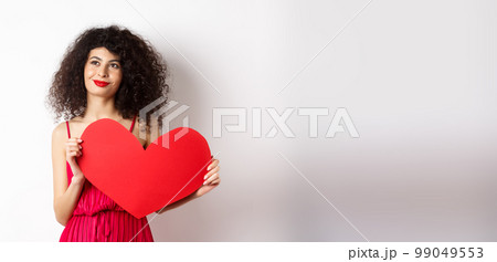 Young romantic woman dreaming about love on Valentines day, looking for soulmate, holding big red heart cutout and gazing pensive left, white background 99049553