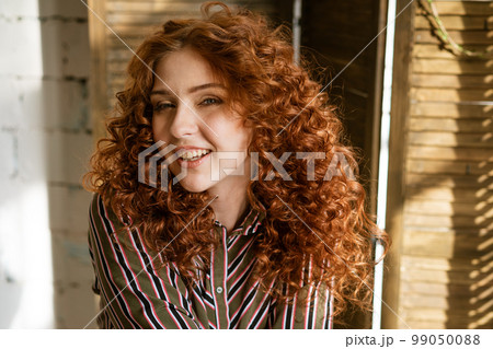 Portrait of happy redhead young woman near window Portrait of happy redhead young woman near window 99050088