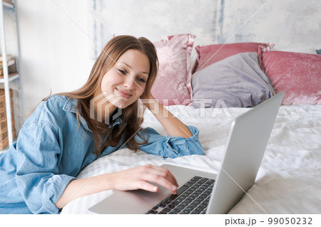Young woman relaxes using laptop on cold winter day in bedroom while sitting Young woman relaxes using laptop on cold winter day in bedroom while sitting 99050232