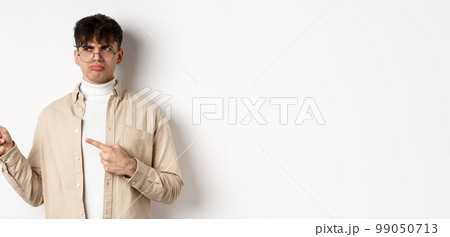 Disappointed guy in glasses looking and pointing left, sulking displeased, complaining on bad offer, standing upset on white background 99050713