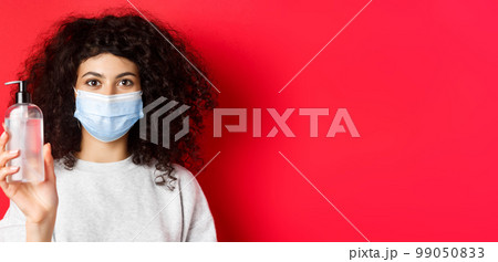 Covid-19, pandemic and quarantine concept. Young woman in medical mask showing bottle of hand sanitizer, demonstrate antiseptic, red background 99050833
