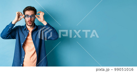 Funny young man trying new sunglasses, looking surprised at camera, standing on blue background Funny young man trying new sunglasses, looking surprised at camera, standing on blue background 99051016