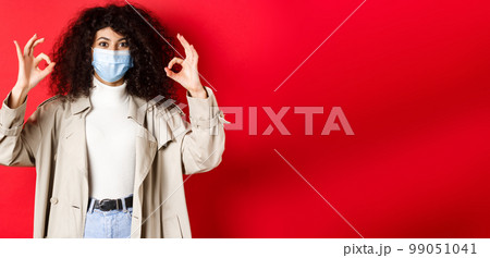 Covid-19, social distancing and quarantine concept. Fashionable woman with curly hair, wearing medical mask and trench coat, showing okay gestures, red background 99051041