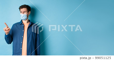 Covid-19 and healthcare concept. Confused and doubtful man in face mask frowning, pointing and looking at empty space, standing on blue background Covid-19 and healthcare concept. Confused and doubtful man in face mask frowning, pointing and looking at empty space, standing on blue background 99051125