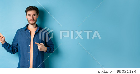 Handsome young man in shirt, pointing fingers left at promo, showing logo, standing on blue background 99051134