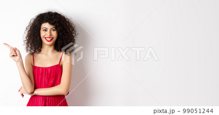 Beautiful happy woman with curly hairstyle, wearing festive dress, pointing finger left at logo and smiling, white background 99051244