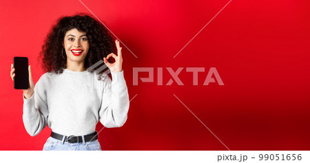 Cheerful european woman with curly hair, showing empty mobile phone screen and okay gesture, smiling satisfied, praise good app or promotion, red background Cheerful european woman with curly hair, showing empty mobile phone screen and okay gesture, smiling satisfied, praise good app or promotion, red background 99051656