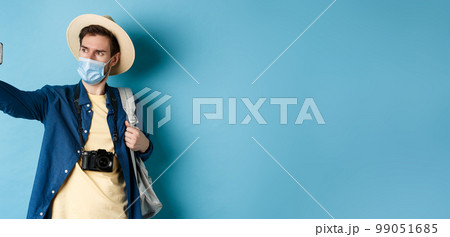 Covid-19, pandemic and travel concept. Tourist on summer vacation taking selfie in medical mask, photographing on smartphone, standing on blue background 99051685
