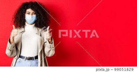 Covid-19, social distancing and quarantine concept. Cheerful stylish woman wearing trench coat and medical mask for going outside during pandemic, showing thumb up 99051829
