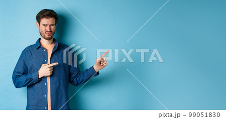 Disappointed young man frowning and pointing fingers right at empty space, looking upset and displeased, complaining, standing on blue background Disappointed young man frowning and pointing fingers right at empty space, looking upset and displeased, complaining, standing on blue background 99051830