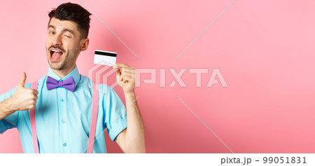 Shopping concept. Festive guy in bow-tie winking at camera, showing thumbs up with plastic credit card, recommending good shop offer, promote bank, standing on pink background Shopping concept. Festive guy in bow-tie winking at camera, showing thumbs up with plastic credit card, recommending good shop offer, promote bank, standing on pink background 99051831