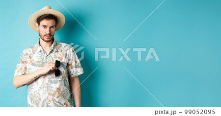 Handsome tourist in hawaiian shirt and summer hat, put sunglasses in pocket, going on vacation, standing against blue background Handsome tourist in hawaiian shirt and summer hat, put sunglasses in pocket, going on vacation, standing against blue background 99052095