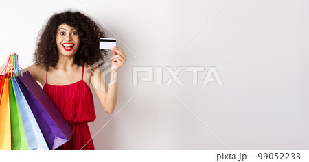 Stylish caucasian woman with curly hair and red dress, showing shopping bags and her plastic credit card, smiling amused, standing over white background Stylish caucasian woman with curly hair and red dress, showing shopping bags and her plastic credit card, smiling amused, standing over white background 99052233