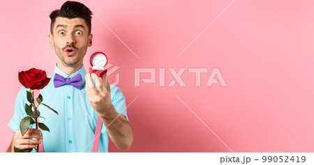Valentines day. Funny man with moustache and bow-tie making proposal, showing engagement ring and propose with red rose, standing over pink background Valentines day. Funny man with moustache and bow-tie making proposal, showing engagement ring and propose with red rose, standing over pink background 99052419