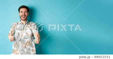 Excited smiling guy in hawaiian shirt show thumbs up, looking amazed, checking out cool promo, standing on blue background Excited smiling guy in hawaiian shirt show thumbs up, looking amazed, checking out cool promo, standing on blue background 99052531