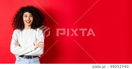 Cheerful young woman with curly hairstyle, raising eyebrows and looking surprised, hear interesting news, red background Cheerful young woman with curly hairstyle, raising eyebrows and looking surprised, hear interesting news, red background 99052540