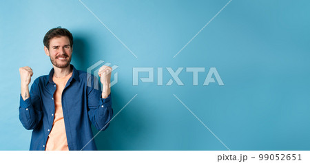 Image of happy man celebrating success, shouting yes and shaking clenched fists, achieve goal and triumphing, winning and dancing, standing on blue background Image of happy man celebrating success, shouting yes and shaking clenched fists, achieve goal and triumphing, winning and dancing, standing on blue background 99052651