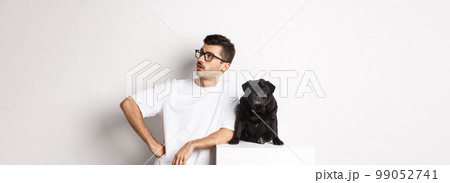 Handsome young man standing near cute black pug, looking right with arrogant expression, standing over white background 99052741