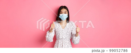 Covid-19, quarantine and lifestyle concept. Excited asian woman in face mask showing thumbs-up, praising good job, looking amazed at camera, standing over pink background Covid-19, quarantine and lifestyle concept. Excited asian woman in face mask showing thumbs-up, praising good job, looking amazed at camera, standing over pink background 99052786
