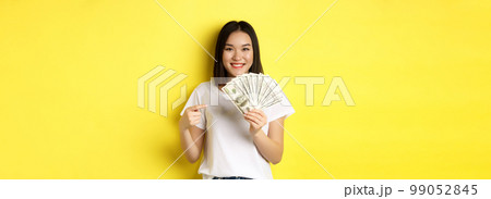 Young asian woman smiling, showing prize money, pointing finger at dollars, standing over yellow background Young asian woman smiling, showing prize money, pointing finger at dollars, standing over yellow background 99052845