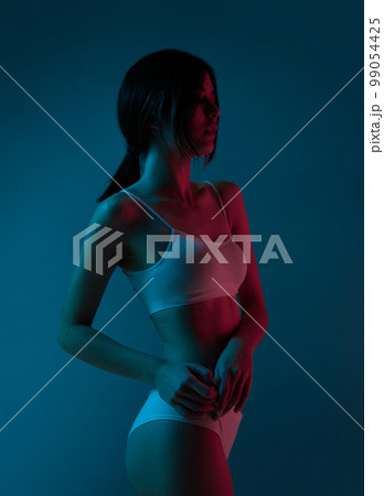 Tenderness. Portrait of beautiful young girl with slim fit body posing in underwear over blue studio background in neon light. Concept of body and skincare, beauty Tenderness. Portrait of beautiful young girl with slim fit body posing in underwear over blue studio background in neon light. Concept of body and skincare, beauty 99054425