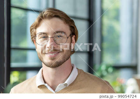 Close-up portrait of a young man inside the office, a businessman in glasses is smiling and looking at the camera, a blond programmer in casual clothes with a beard. 99054724