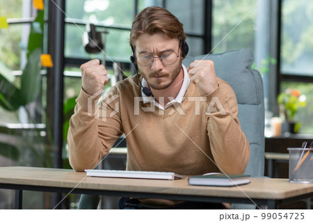 Angry office worker working inside office, young businessman in casual clothes with headset for video call, male nervous tech support worker and customer consultation online. 99054725