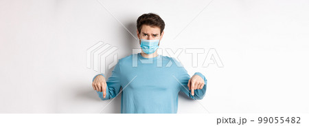 Covid-19, health and quarantine concept. Sad man complaining and looking disappointed, pointing fingers down at logo, standing in medical mask from coronavirus pandemic 99055482