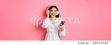 Beautiful asian woman listening music in headphones, using mobile phone, smiling happy at camera, standing over pink background 99055490